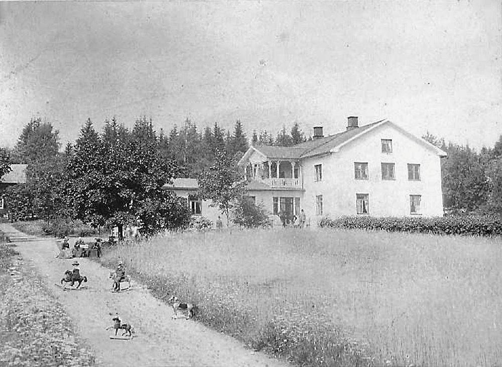Sommaridyll 1895, med barn på varsin gunghäst på grusvägen och tre damer i hatt i varsin vilstol.