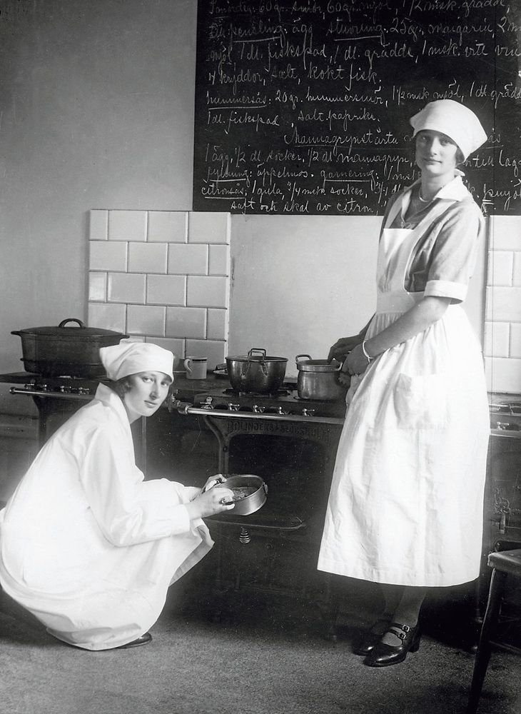 1920-tal. Kronprinsessan Märtha och hennes syster Astrid i ett kök med recept på en stor tavla ovanför spisen.